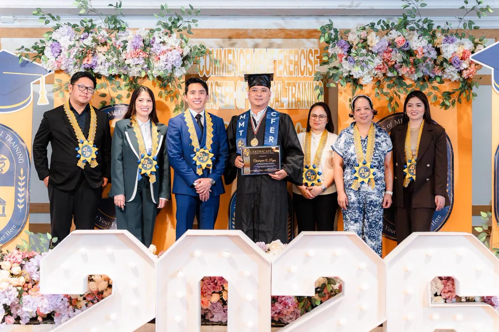 MMT commencement exercise 2026 — graduate holding a certificate with medallion-bearers beside